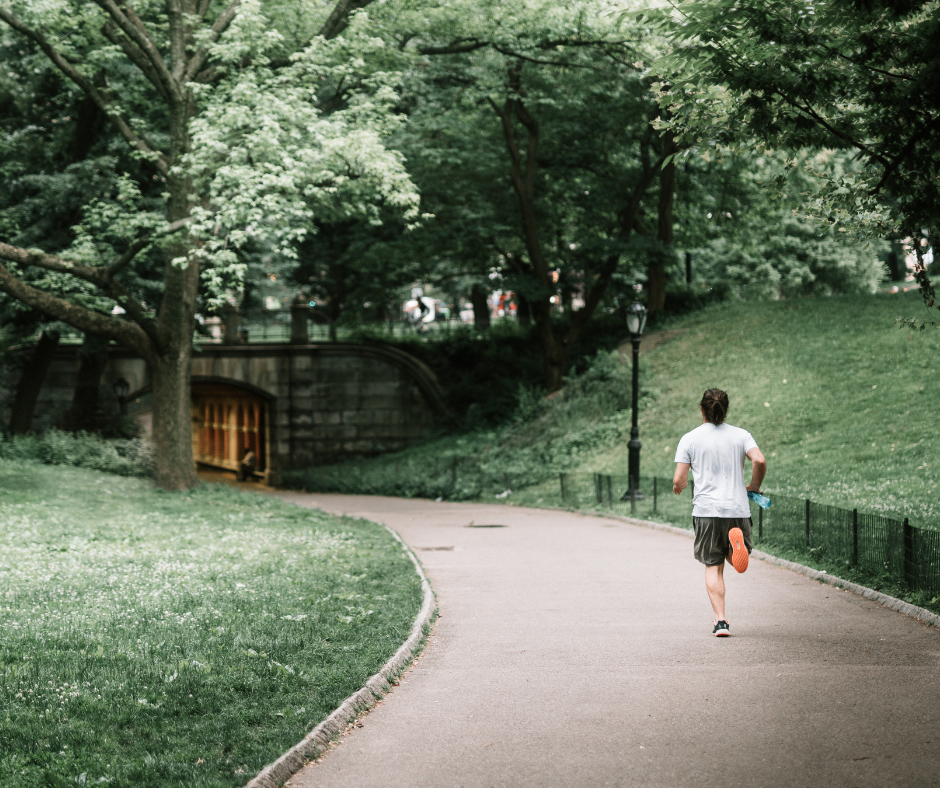 A man jogging down an asphalt path through a park with grass and trees