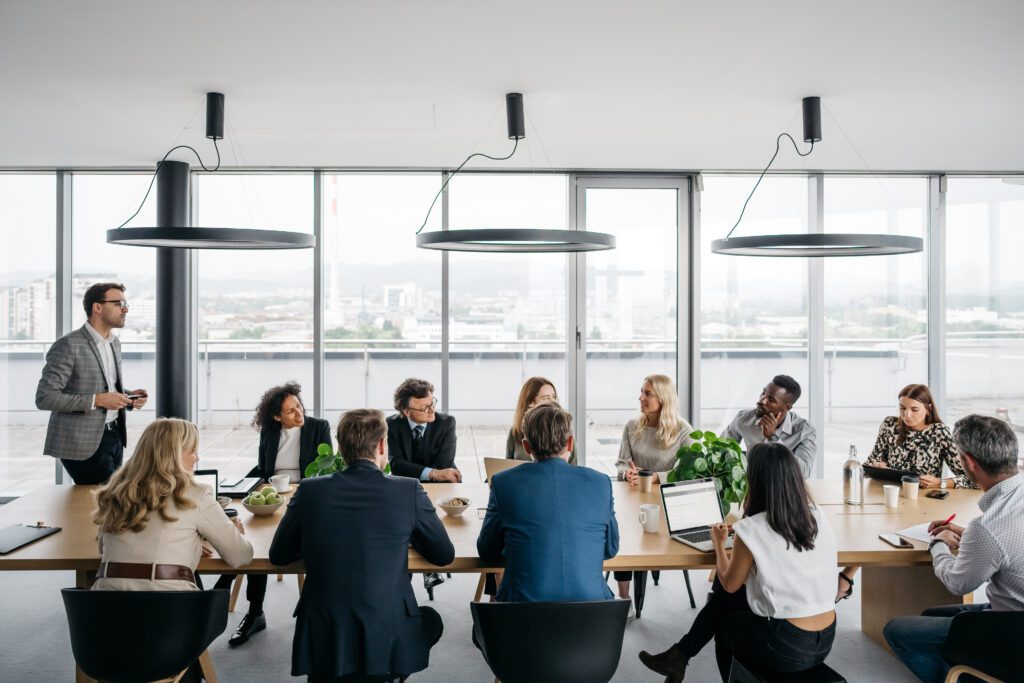 A business meeting around a large boardroom table in a bright office with large windows. A man is standing up while his colleagues are sitting down.